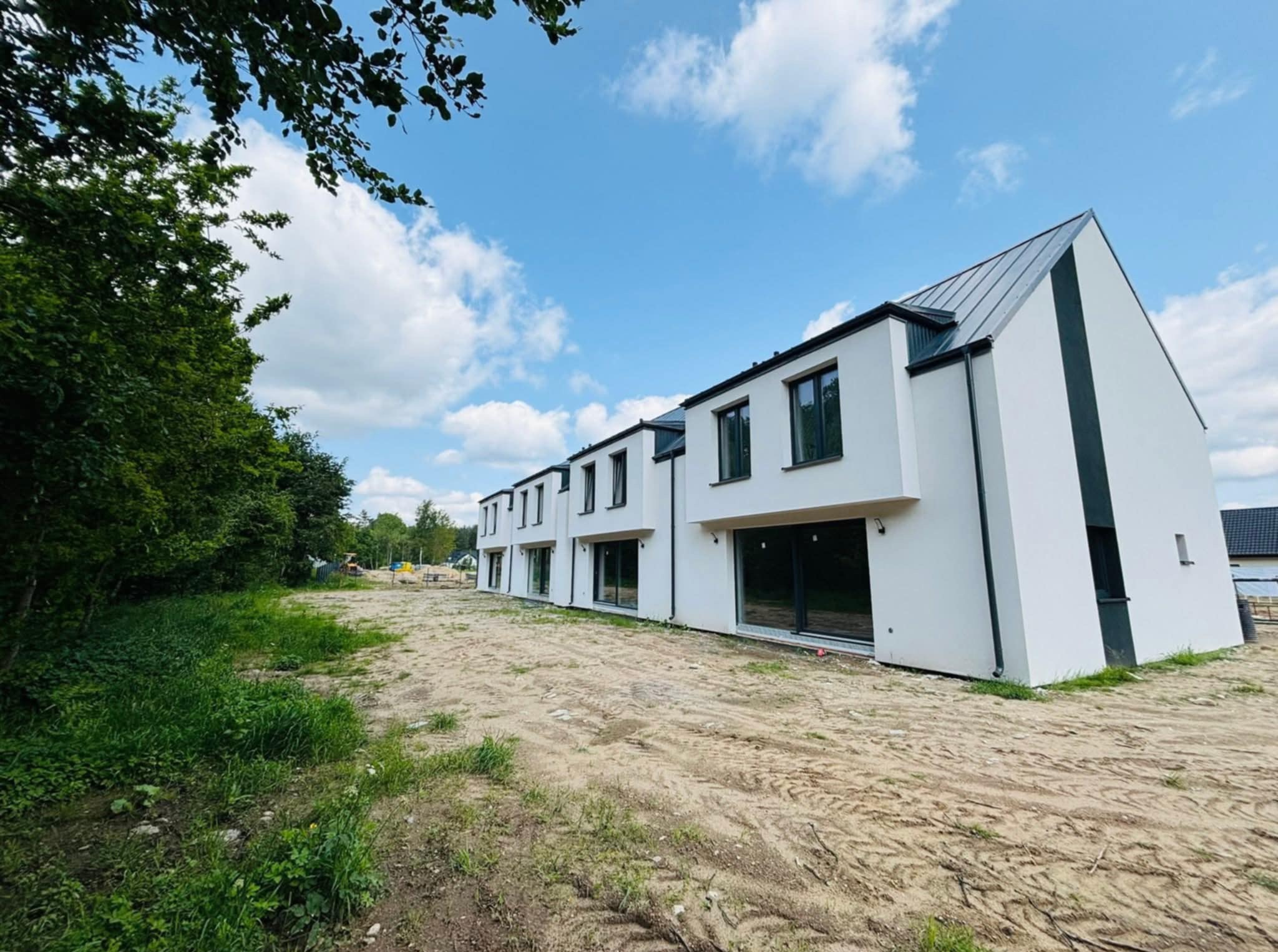 Nowoczesny budynek szeregowy z białą elewacją i czarnymi detalami, w naturalnym otoczeniu z drzewami i błękitnym niebem. Widok z perspektywy trawnika.