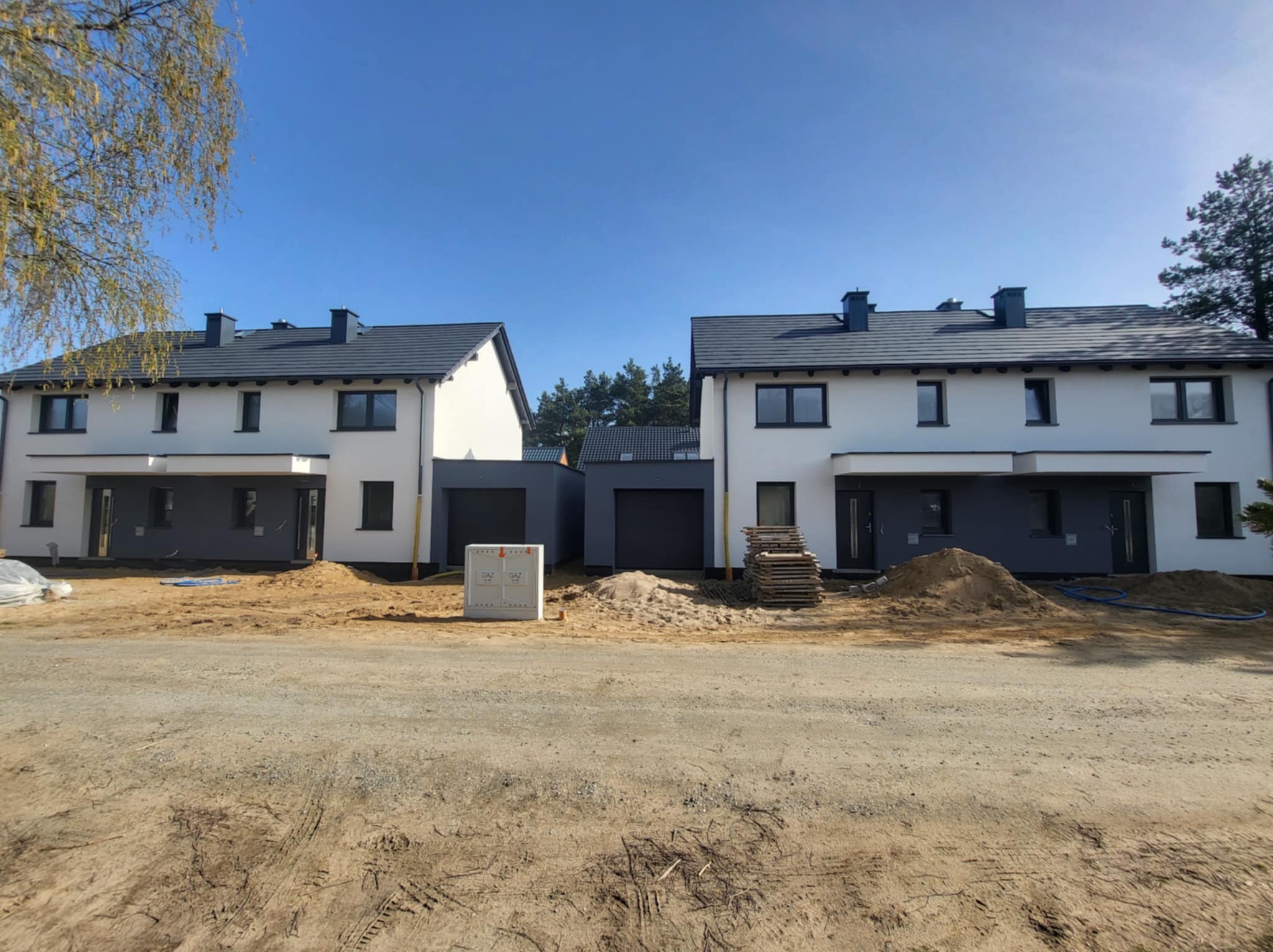 Dwa bliźniacze domy z szarą stolarką i białymi elewacjami, na tle błękitnego nieba. Widoczne elementy wykończeniowe i teren przed budynkami w trakcie prac.