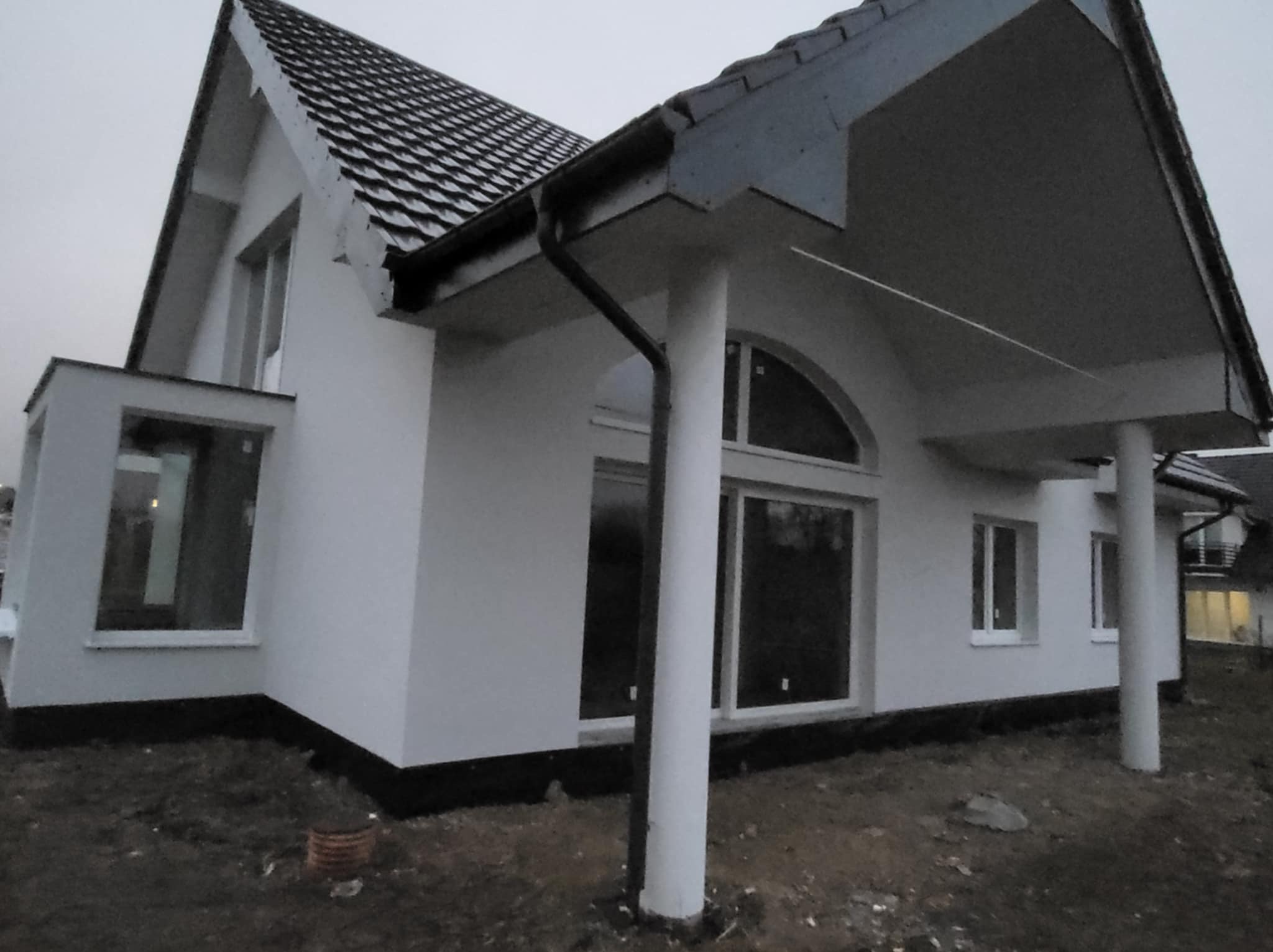 Nowoczesny dom jednorodzinny z białą elewacją, widoczne duże okna i kolumny podtrzymujące zadaszenie. Dom w trakcie wykończenia, teren wokół jeszcze niezagospodarowany.