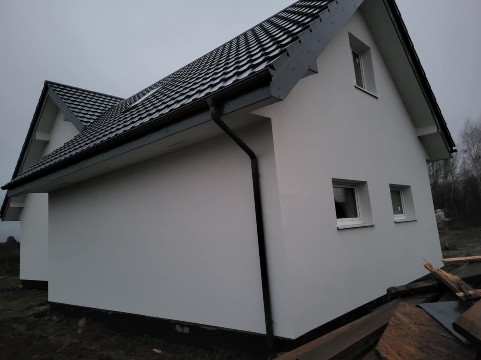 Boczny widok domu z białą elewacją i ciemną dachówką. Widoczne okna, rynny i fragment terenu wokół budynku. Pochmurne niebo w tle.