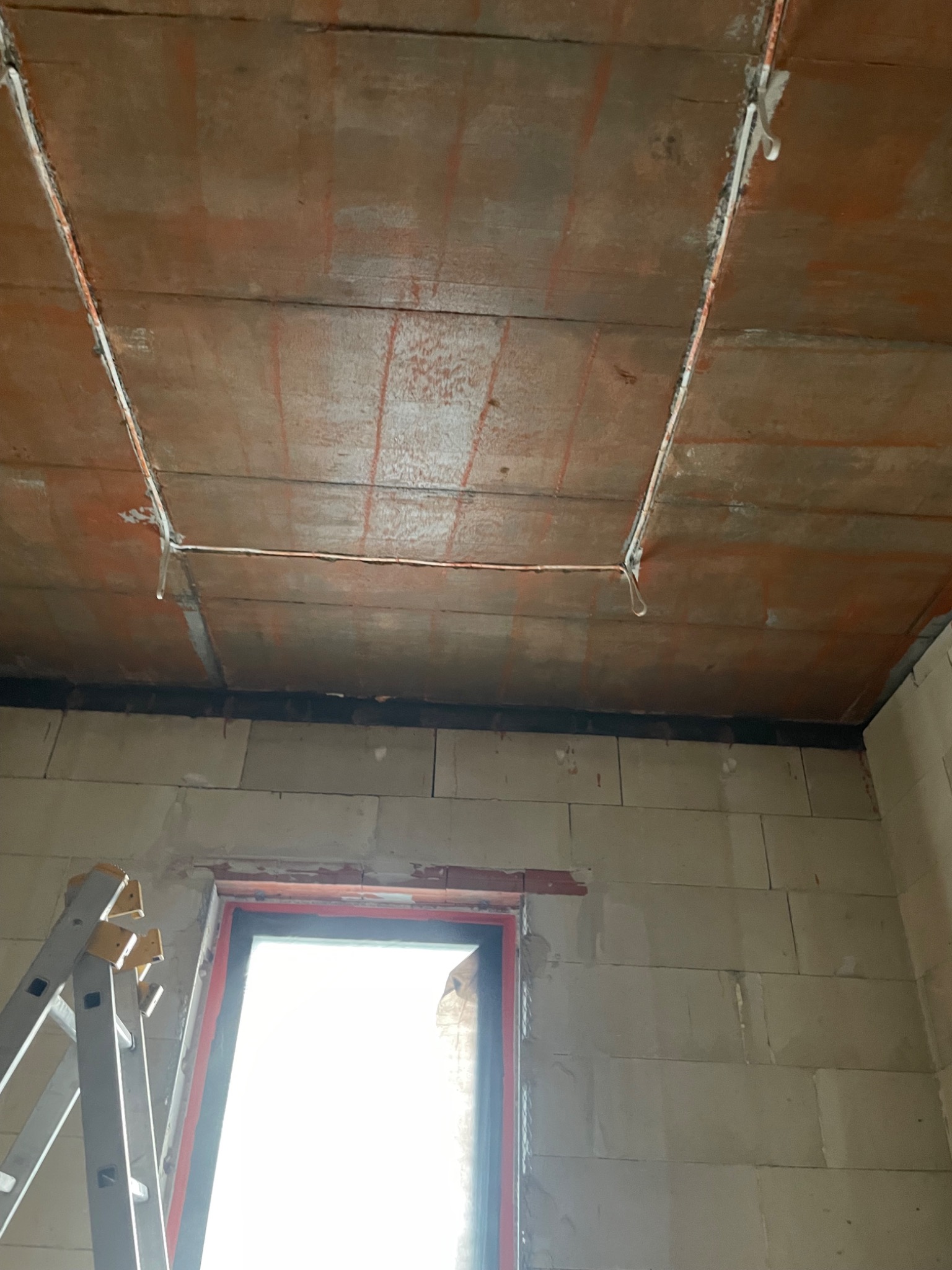 Montaż przewodów elektrycznych pod sufitem z pustaków, widoczna drabina i okno. Instalacja w trakcie budowy, surowy stan.