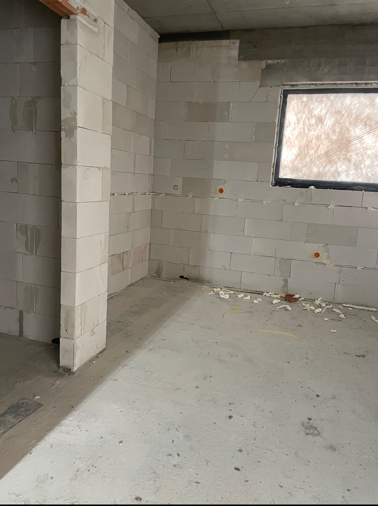Surowy stan wnętrza z pustaków, widoczne punkty instalacji elektrycznej, okno zabezpieczone folią, resztki materiałów budowlanych na posadzce.