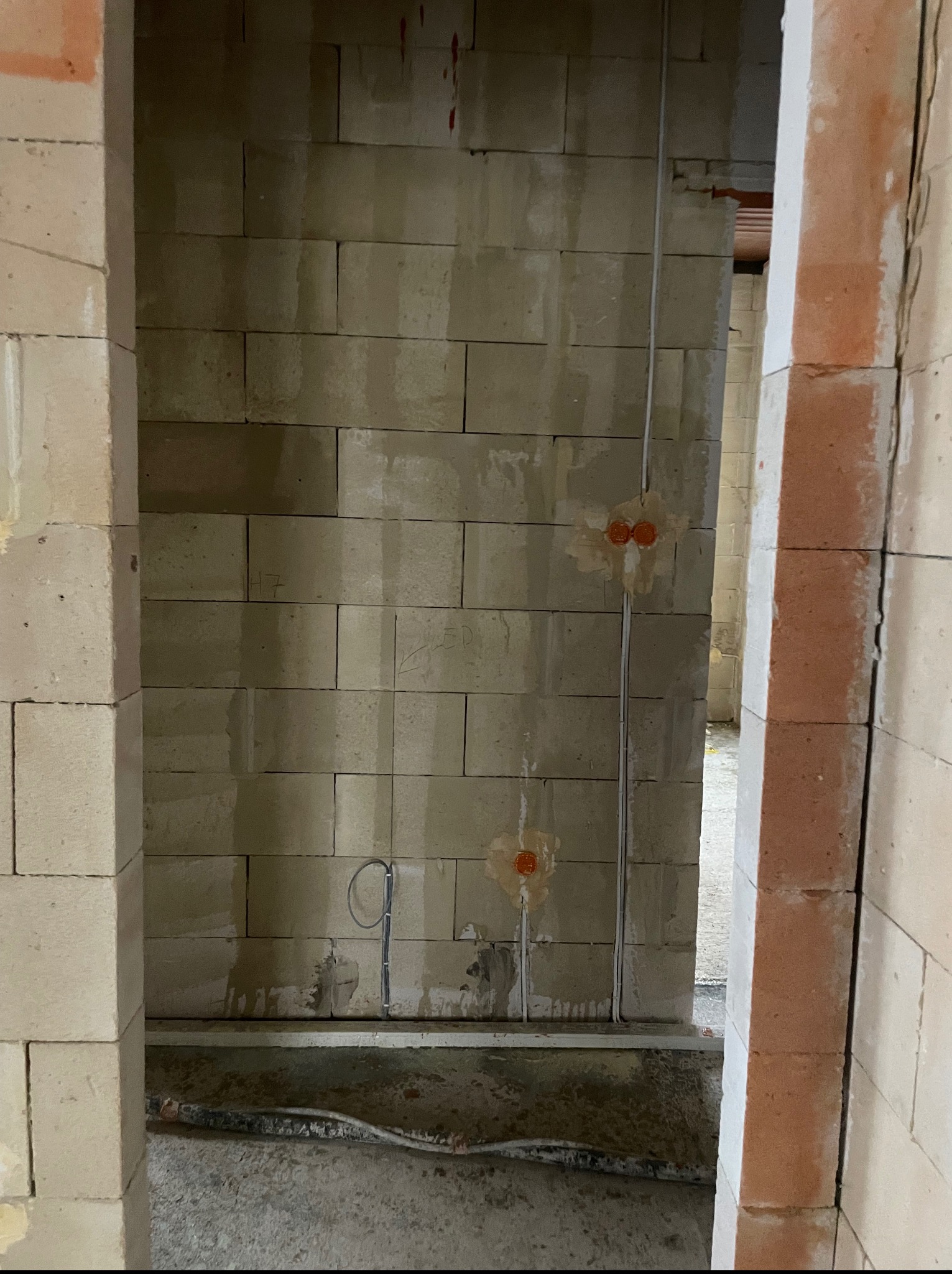 Surowa ściana z betonu komórkowego z widocznymi przewodami elektrycznymi i puszkami instalacyjnymi, w trakcie prac wykończeniowych. Kątowe ujęcie z perspektywy wnętrza.