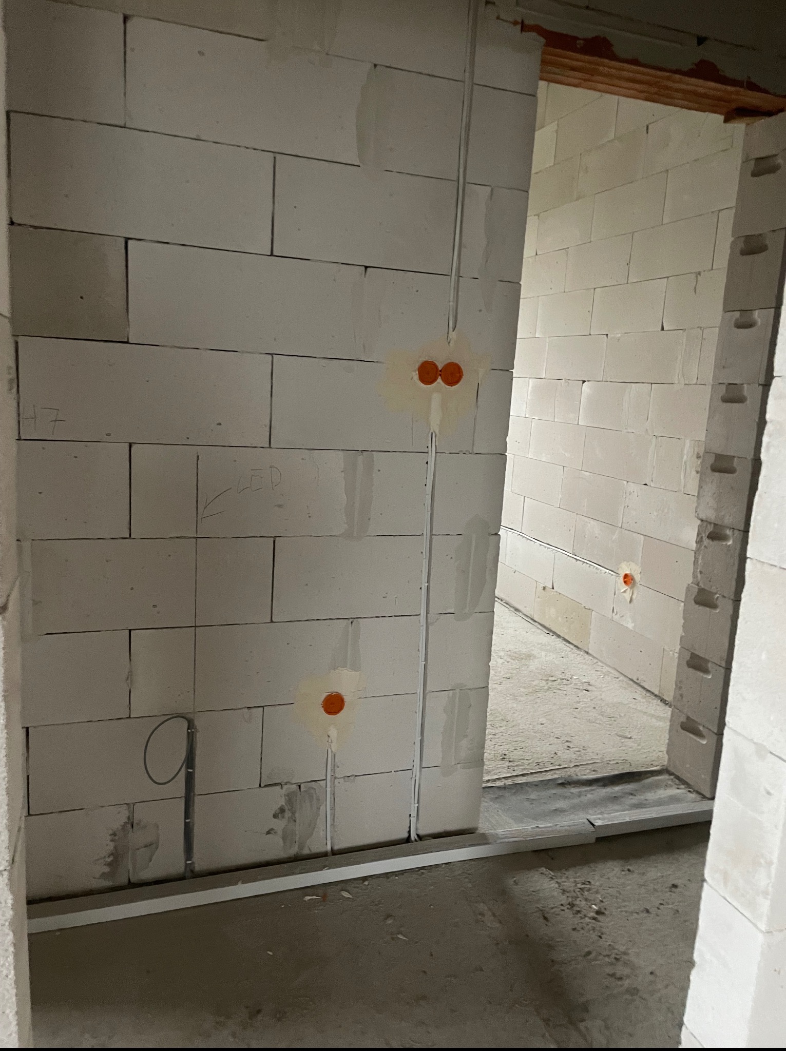 Wewnętrzna ściana z betonu komórkowego w trakcie budowy z widocznymi przewodami elektrycznymi w peszlach. Otwory pod gniazdka zaznaczone pomarańczową farbą.
