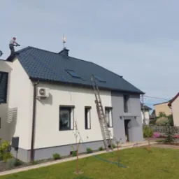 Pracownik w uprzęży czyści ciemnoszary dach nowoczesnego domu z drabiny. Widoczne okna dachowe, antena, fragment elewacji i zadbany ogród.