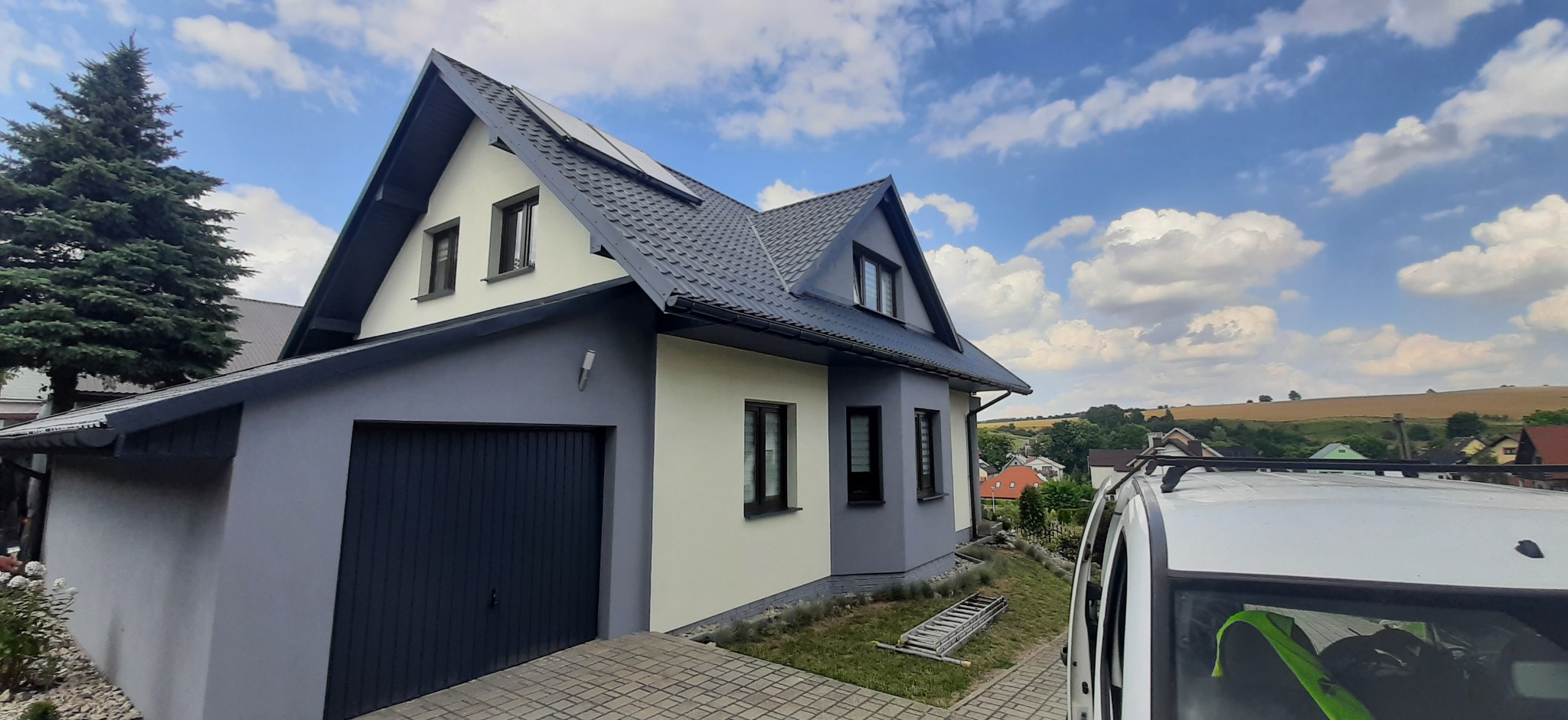 Dom jednorodzinny po renowacji elewacji i dachu, z ciemnoszarą dachówką i szarą elewacją. Na trawniku leży drabina. Błękitne niebo z chmurami w tle.