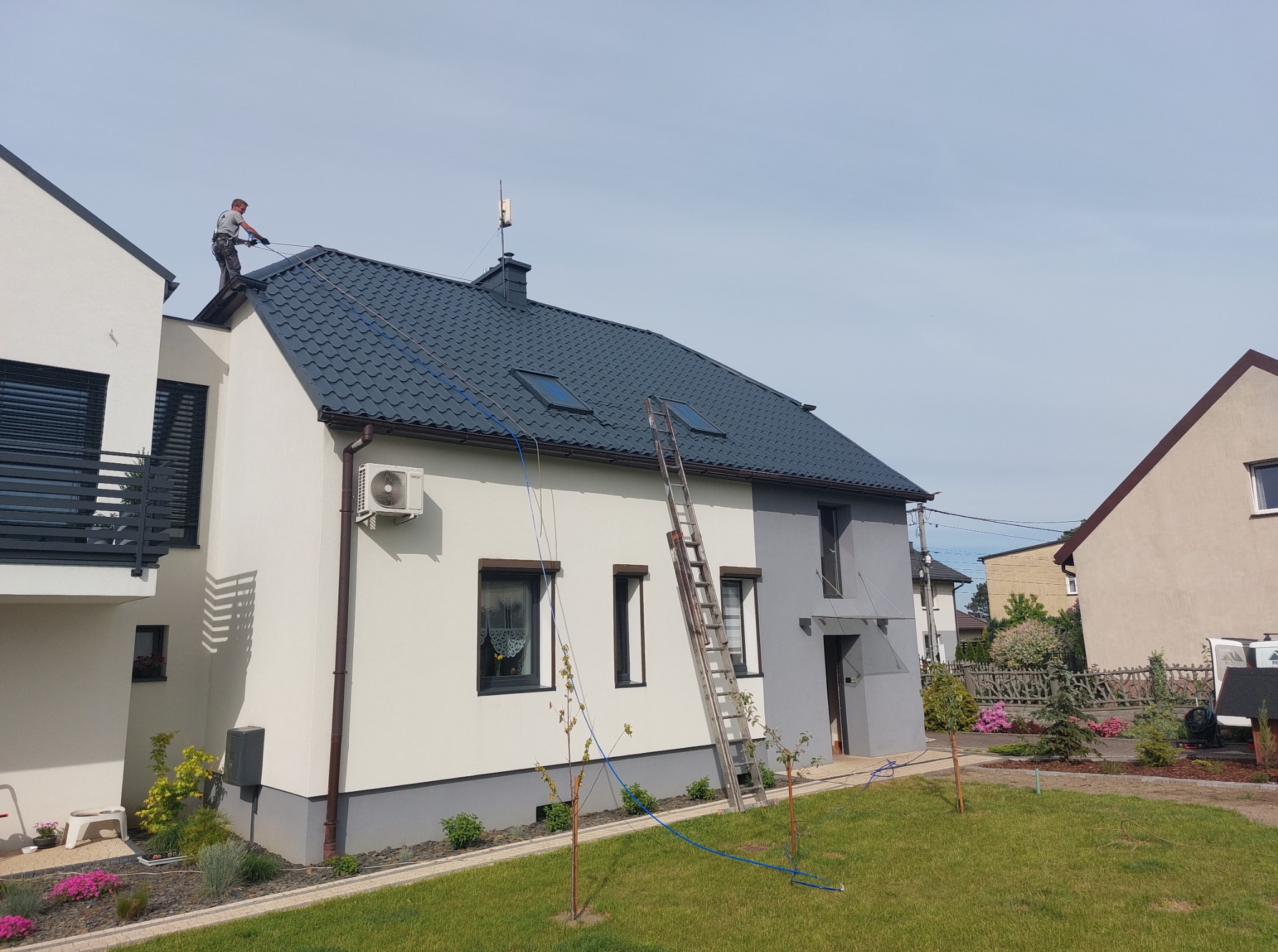 Pracownik w uprzęży czyści ciemnoszary dach nowoczesnego domu z drabiny. Widoczne okna dachowe, antena, fragment elewacji i zadbany ogród.