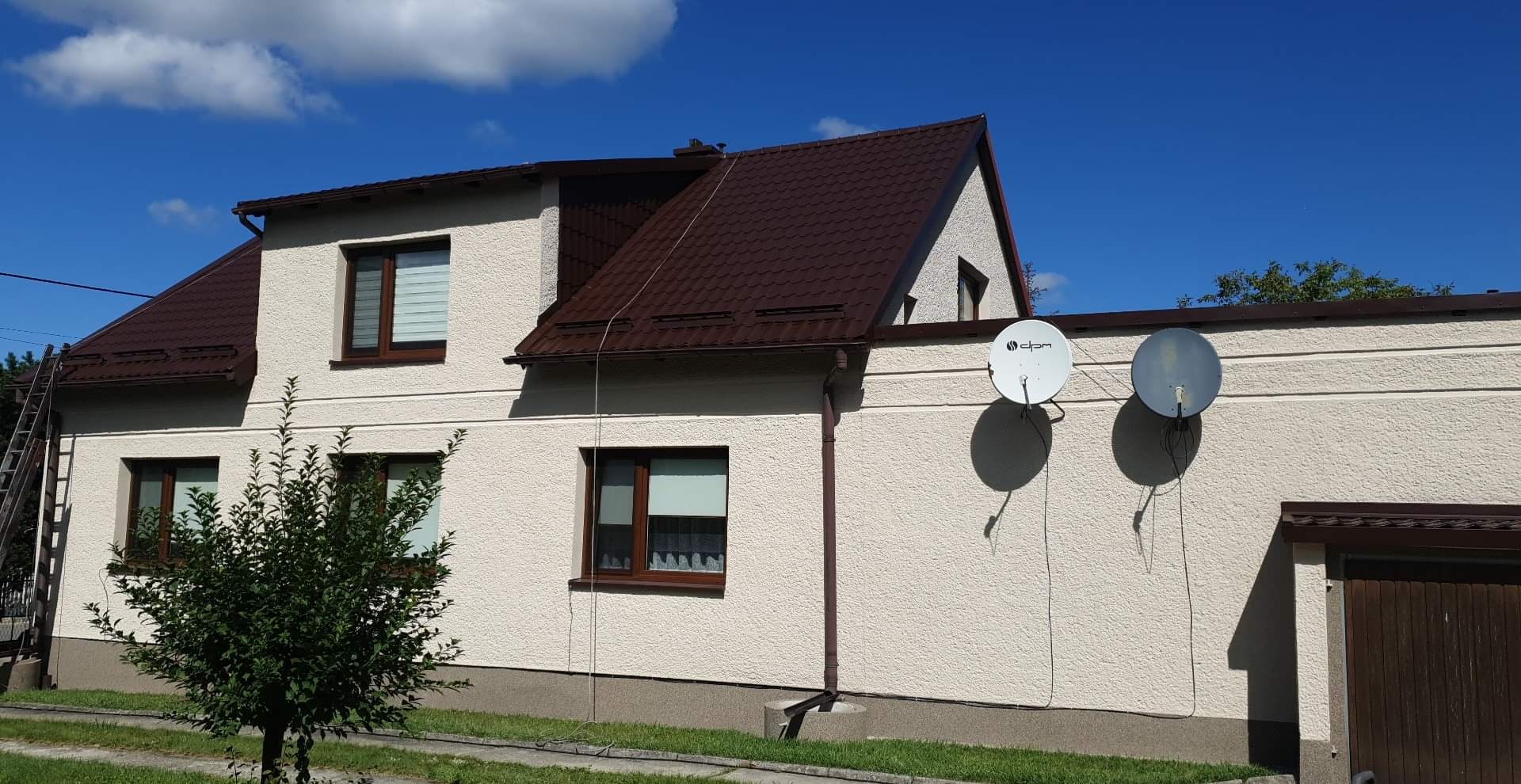 Dwukondygnacyjny dom z brązowym dachem i beżową elewacją w słoneczny dzień. Widoczne dwie anteny satelitarne i drabina oparta o ścianę. Mały krzew na pierwszym planie.