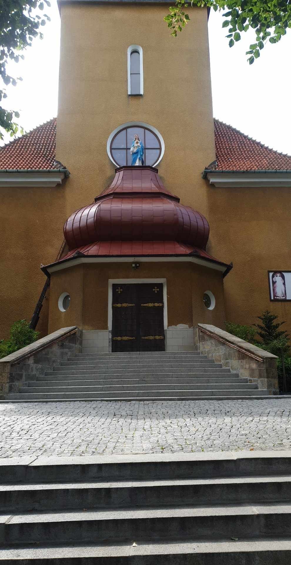 Kościół z beżową elewacją, czerwonym dachem kopułowym i schodami z kostki brukowej. Nad wejściem okrągłe okno z figurą Matki Boskiej.