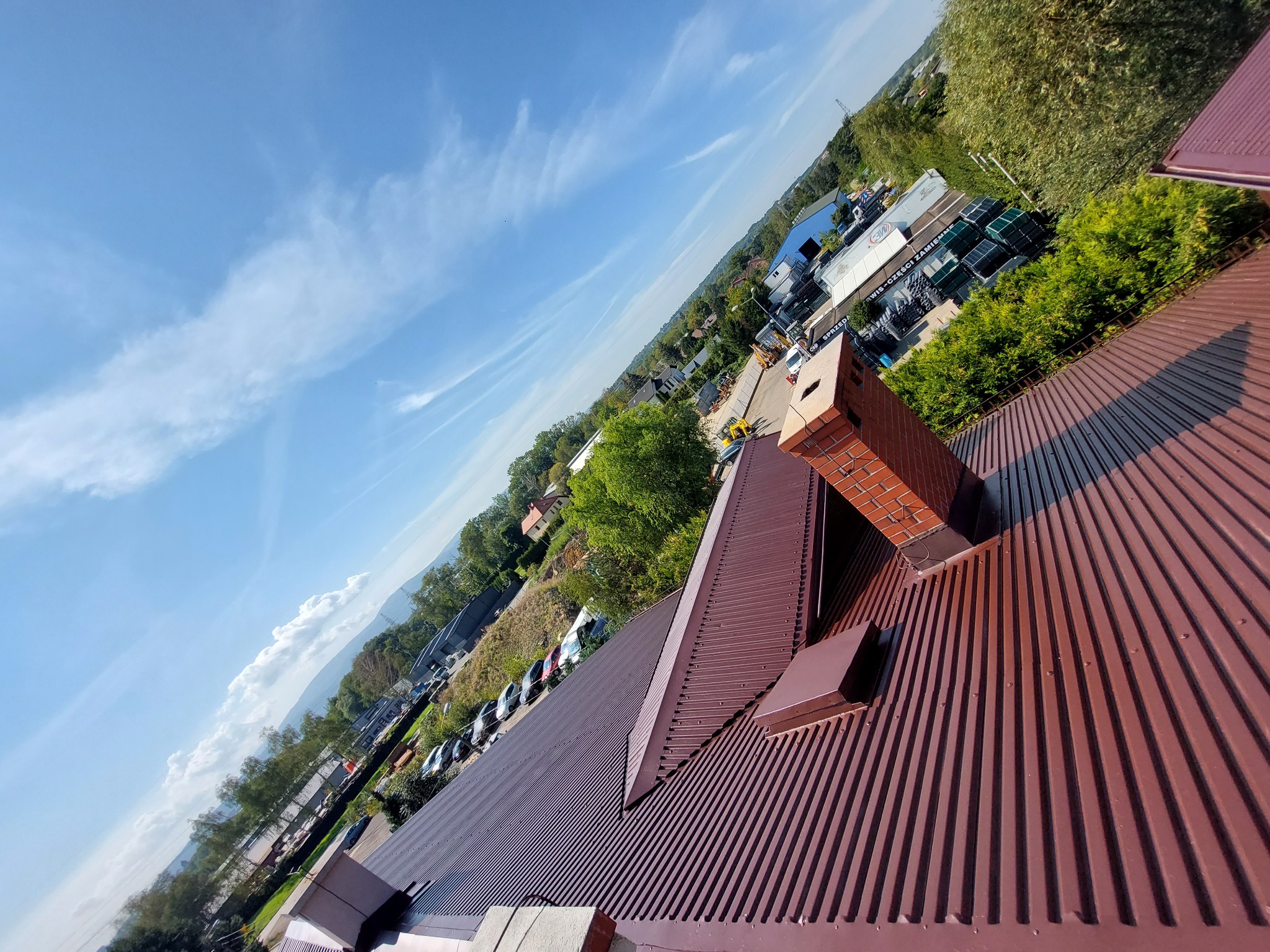 Czerwono-brązowy dach z blachy trapezowej, ceglany komin, błękitne niebo z chmurami w tle. Ujęcie z góry, ukośne. Widok na okolicę.