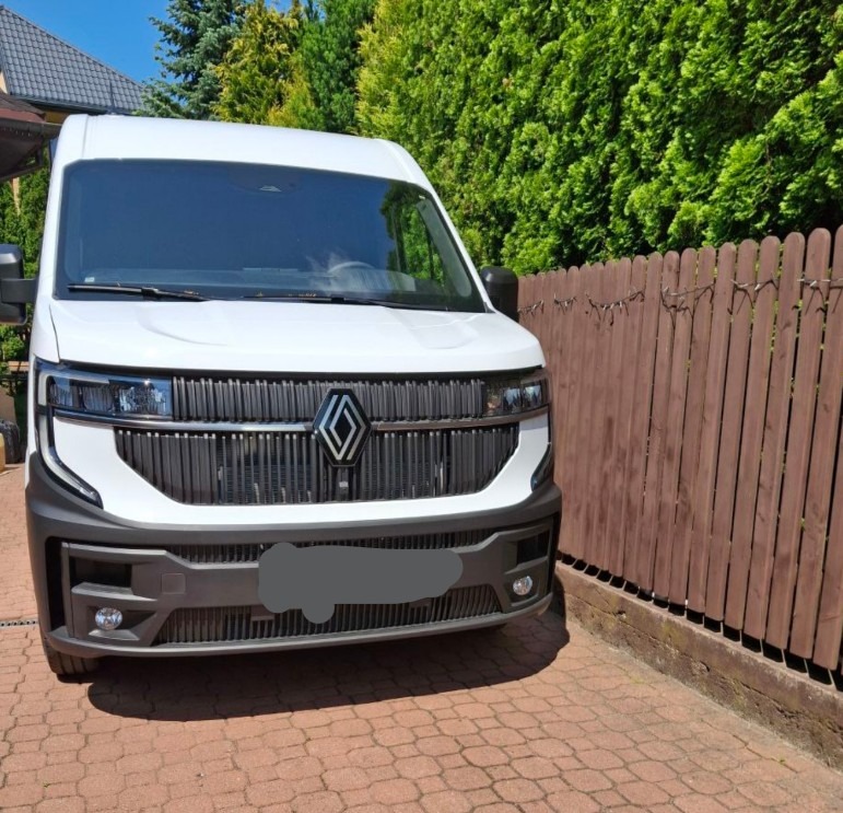Nowoczesny biały bus Renault z przodu, zaparkowany na kostce brukowej, widoczny na tle zielonego żywopłotu i drewnianego płotu. Charakterystyczna atrapa chłodnicy.