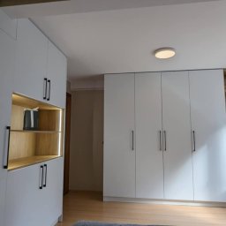 GraStol - Białe szafy na wymiar z czarnymi uchwytami i drewnianymi wstawkami z oświetleniem LED, dopasowane do skosów poddasza. Obok okna regał z książkami.