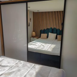 GraStol - Szafa wnękowa z przesuwnymi, białymi drzwiami, odbijająca wnętrze sypialni z drewnianym panelem ściennym za łóżkiem. Minimalistyczny design, nowoczesne wykończenie.