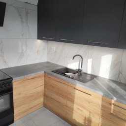 GraStol - Nowoczesna kuchnia: grafitowe szafki górne, drewnopodobne dolne z szarym blatem. Marmurowe płytki na ścianie i podłodze, minimalistyczny zlewozmywak z baterią.