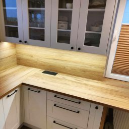 GraStol - Jasna kuchnia z drewnianym blatem i szafkami z przeszkleniami. Nowoczesne czarne uchwyty. Widoczny fragment okna z drewnianymi żaluzjami.