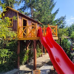 GraStol - Drewniany domek na drzewie z czerwoną zjeżdżalnią w ogrodzie, otoczony zielenią. Konstrukcja wsparta na palach, z żółtymi balustradami. Tabliczka 'Teren Budowy' w tle.