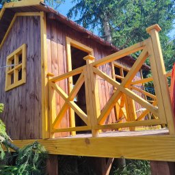 GraStol - Drewniany domek na drzewie z żółtą balustradą i czerwoną zjeżdżalnią, otoczony zielenią drzew, widziany z dołu pod kątem, w słoneczny dzień.