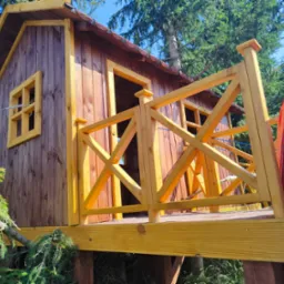 Drewniany domek na drzewie z żółtą balustradą i czerwoną zjeżdżalnią, otoczony zielenią drzew, widziany z dołu pod kątem, w słoneczny dzień.