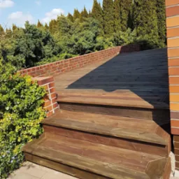 Drewniany taras z trzema schodkami, otoczony zielenią i ceglanym murem, w słoneczny dzień. Widoczne deski tarasowe i elementy konstrukcyjne.