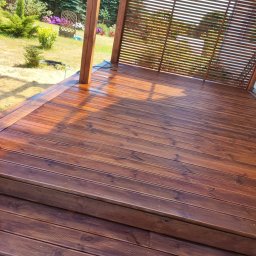 GraStol - Drewniany taras z zadaszeniem lamelowym w Dąbrówce, wykończony w ciepłym, brązowym odcieniu. Widoczne schody i otaczający ogród.