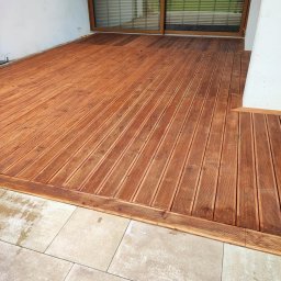 GraStol - Rustykalny taras drewniany z desek ułożonych pod kątem, z obrzeżem i widoczną kostką brukową oraz oknem tarasowym w tle.