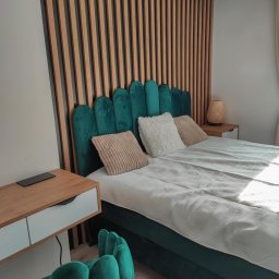 GraStol - Nowoczesna sypialnia z zielonym, aksamitnym łóżkiem i drewnianą ścianą z lameli. Na szafce nocnej stoi lampka. Widoczny fragment zielonego krzesła.