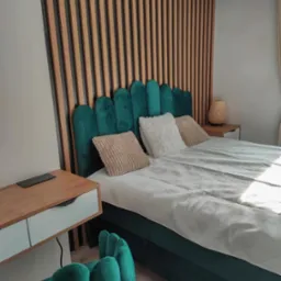 Nowoczesna sypialnia z zielonym, aksamitnym łóżkiem i drewnianą ścianą z lameli. Na szafce nocnej stoi lampka. Widoczny fragment zielonego krzesła.