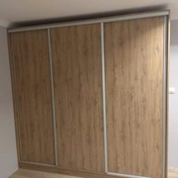 GraStol - Trzydrzwiowa szafa wnękowa z przesuwnymi drzwiami o teksturze jasnego drewna, z subtelnymi srebrnymi obramowaniami, wkomponowana w jasne wnętrze.