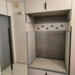 Funkcjonalna zabudowa przedpokoju z lustrem i tapicerowanym siedziskiem z wieszakami. Minimalistyczny design, jasna kolorystyka, czarne uchwyty. Widoczny fragment instalacji elektrycznej.