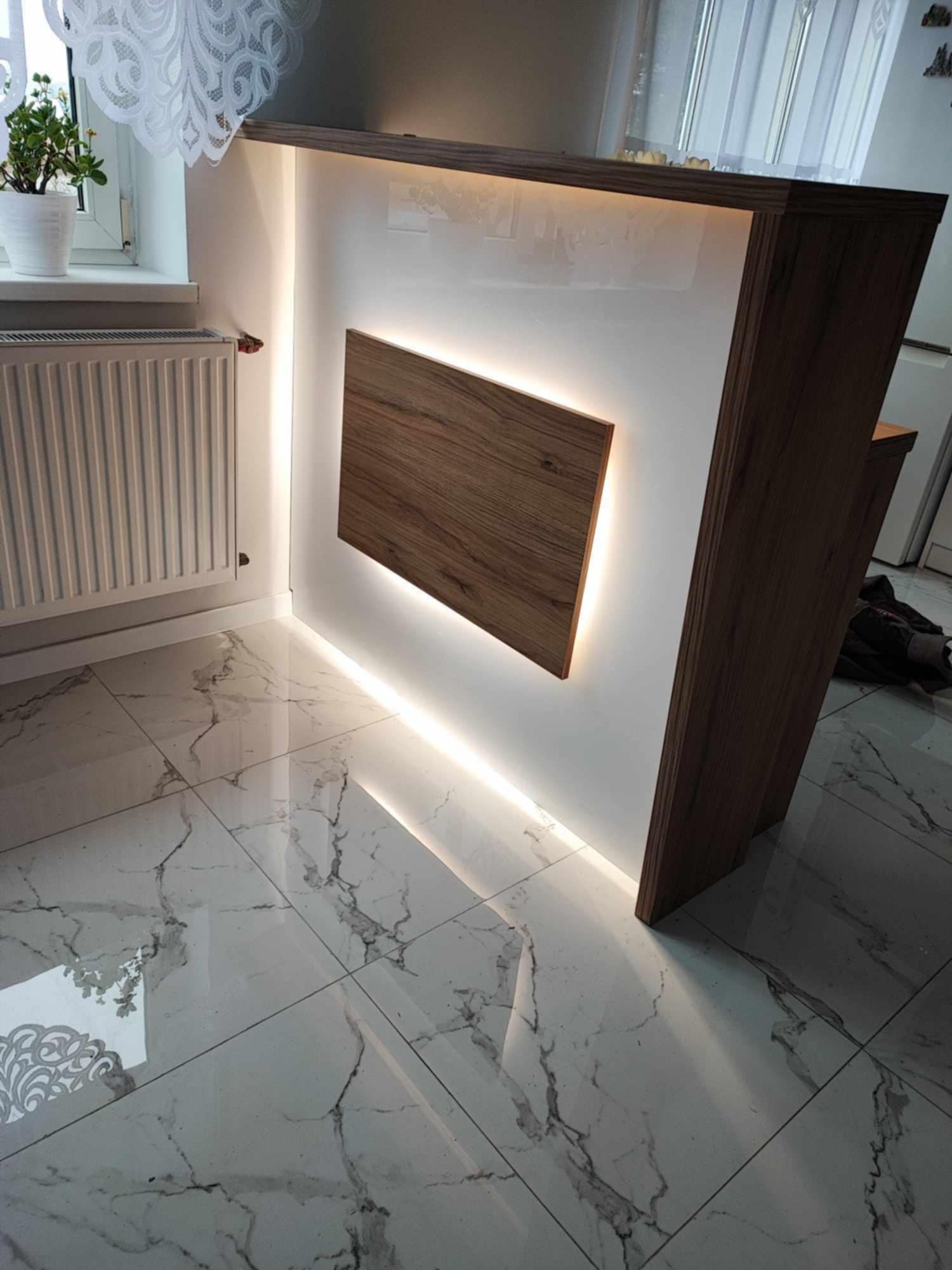 Nowoczesna zabudowa z podświetleniem LED, białe fronty i drewniane akcenty, marmurowe płytki podłogowe, widok z dołu, roślina w oknie.