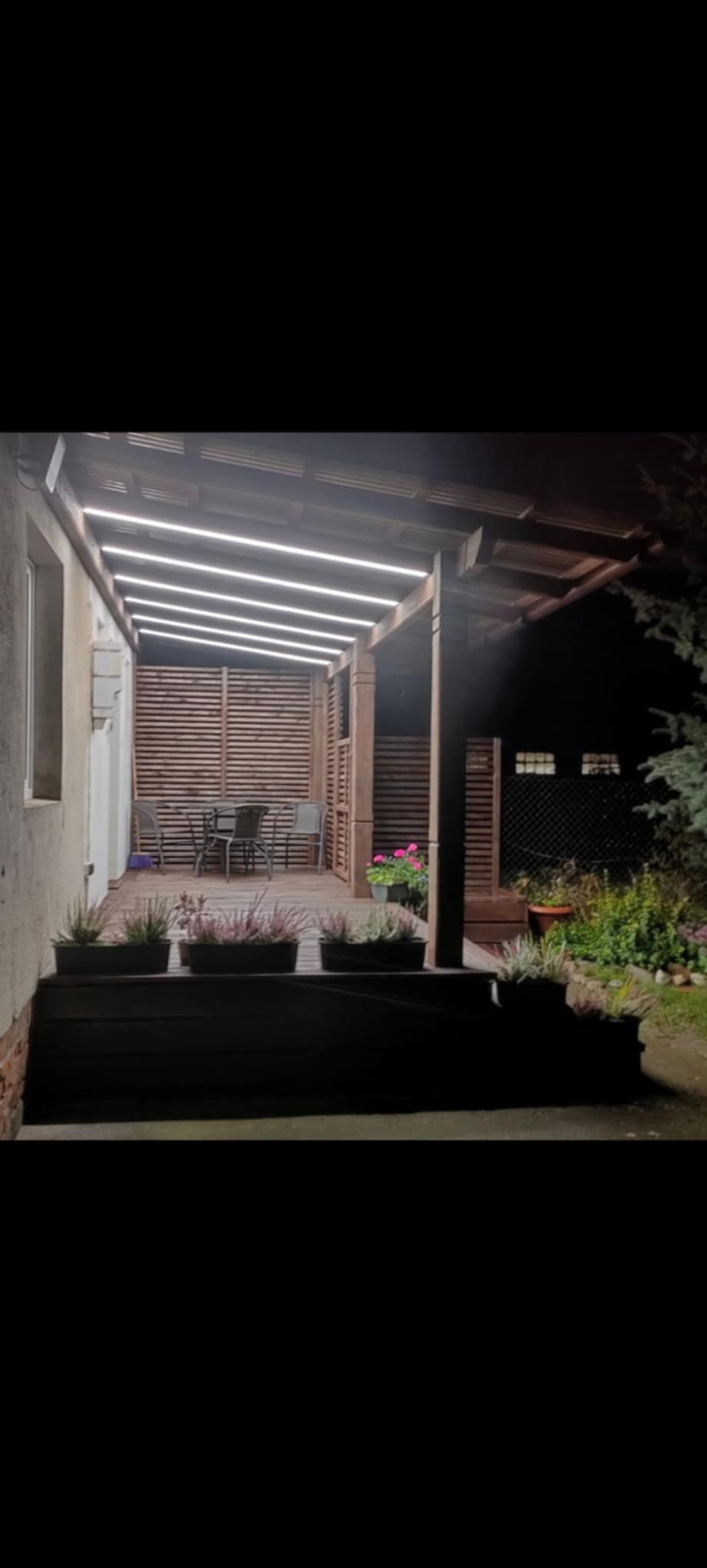 Oświetlony taras drewniany z pergolą i lamelami w tle. Stolik z krzesłami. Donice z roślinami. Nocne oświetlenie LED. Ujęcie z dołu.