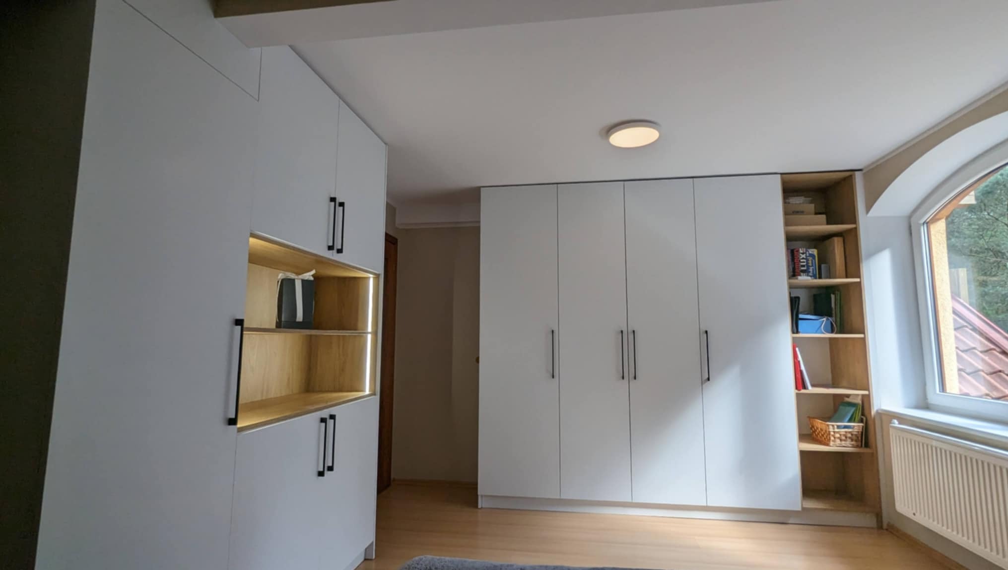 Białe szafy na wymiar z czarnymi uchwytami i drewnianymi wstawkami z oświetleniem LED, dopasowane do skosów poddasza. Obok okna regał z książkami.