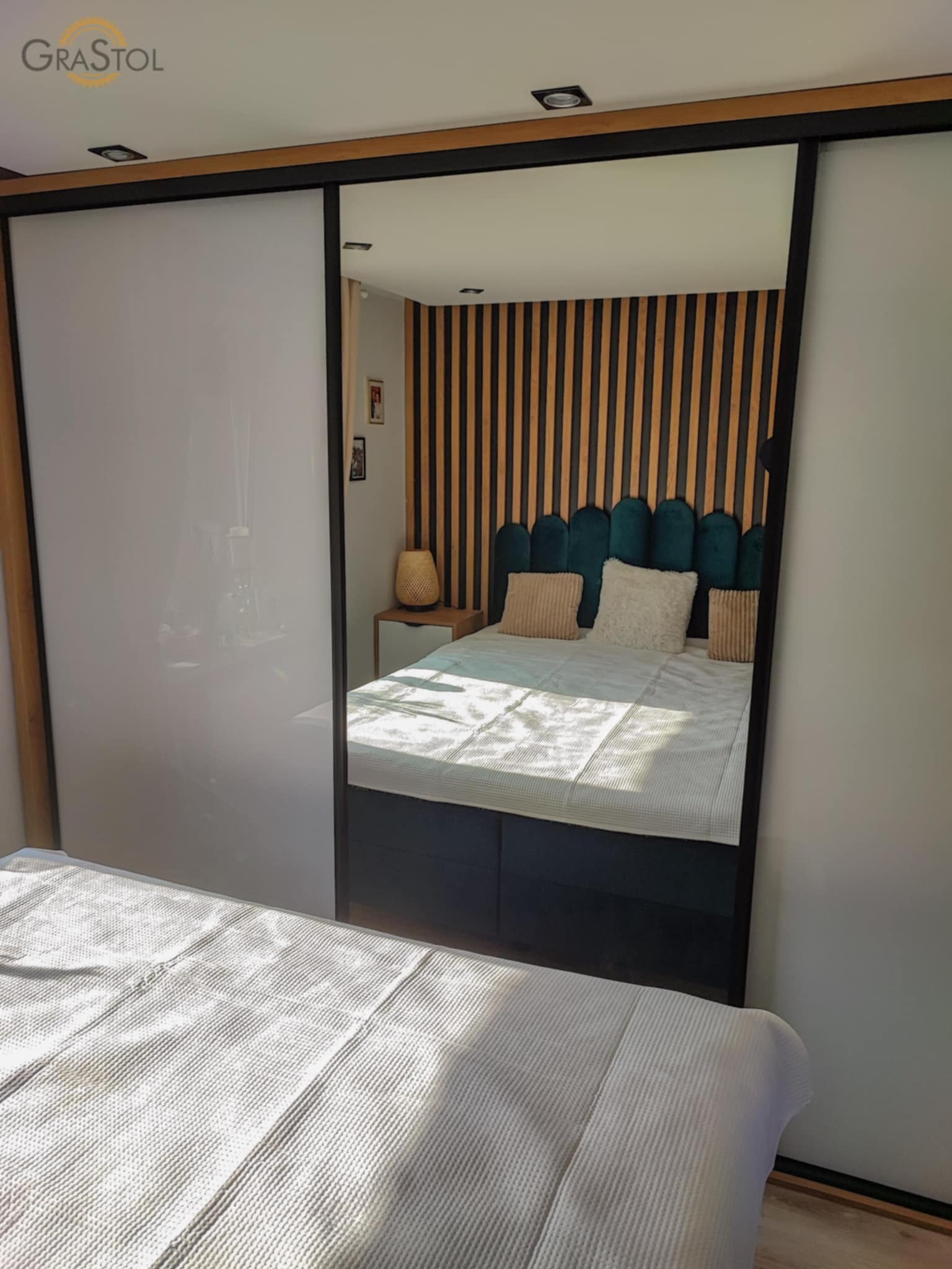 Szafa wnękowa z przesuwnymi, białymi drzwiami, odbijająca wnętrze sypialni z drewnianym panelem ściennym za łóżkiem. Minimalistyczny design, nowoczesne wykończenie.