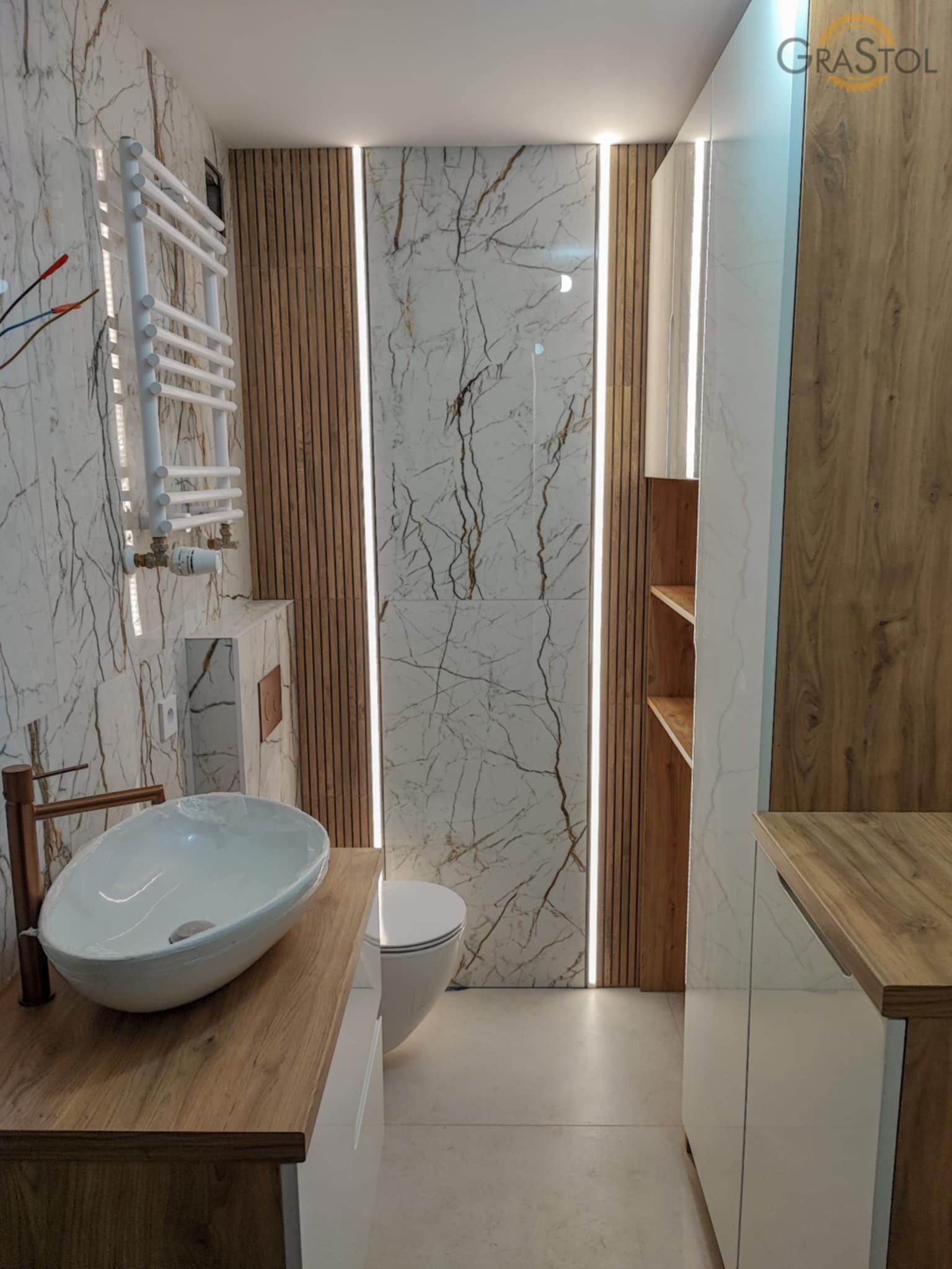 Nowoczesna łazienka z marmurowymi płytkami, drewnianymi akcentami i owalną umywalką na blacie. Grzejnik drabinkowy i oświetlenie LED dodają elegancji.