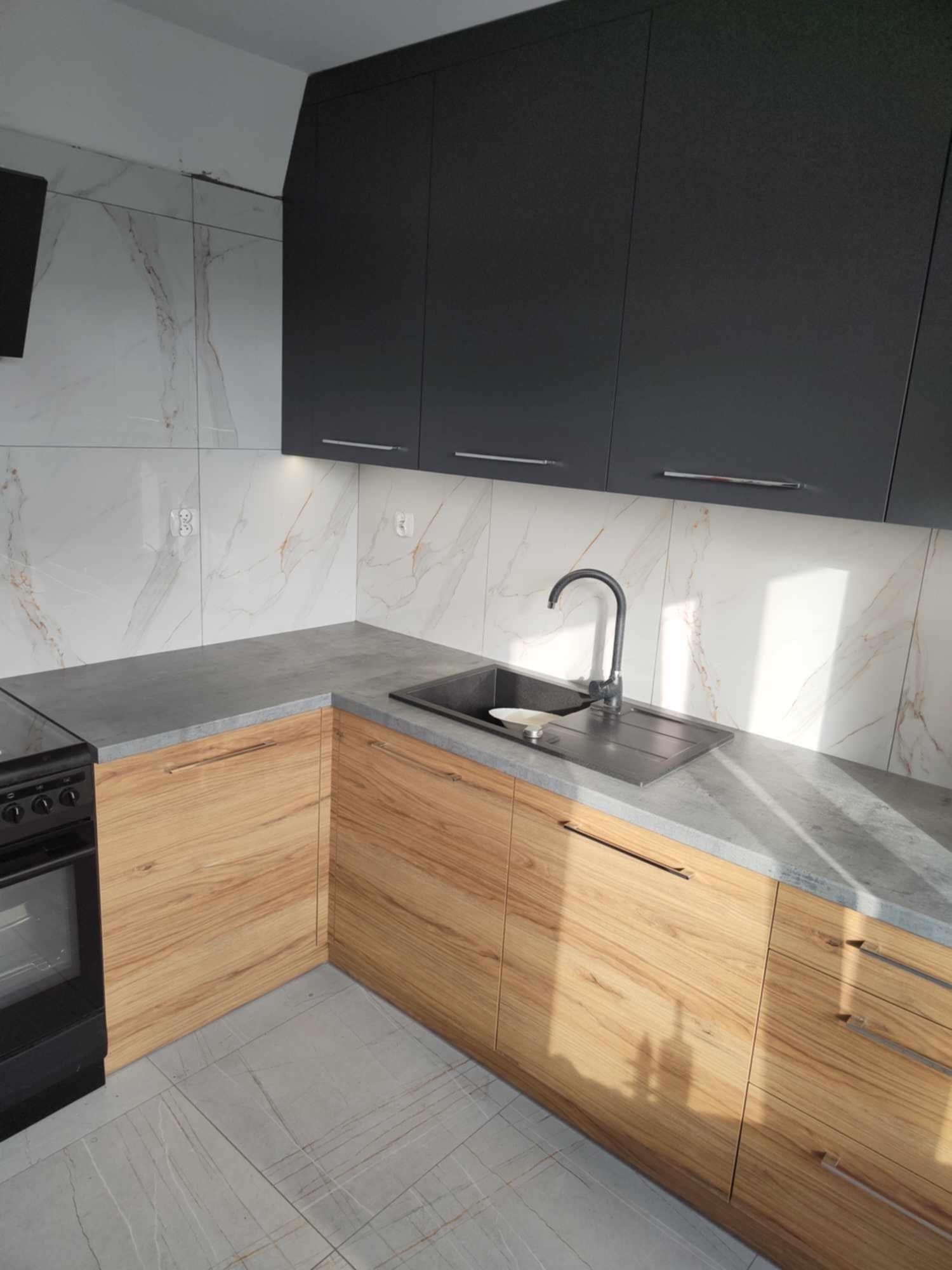 Nowoczesna kuchnia: grafitowe szafki górne, drewnopodobne dolne z szarym blatem. Marmurowe płytki na ścianie i podłodze, minimalistyczny zlewozmywak z baterią.