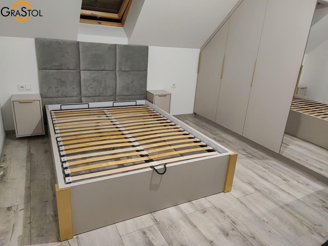 Jasna sypialnia z łóżkiem z szarym wezgłowiem, szafkami nocnymi i szafą wnękową z lustrzanymi drzwiami. Widoczna drewniana podłoga i okno dachowe.