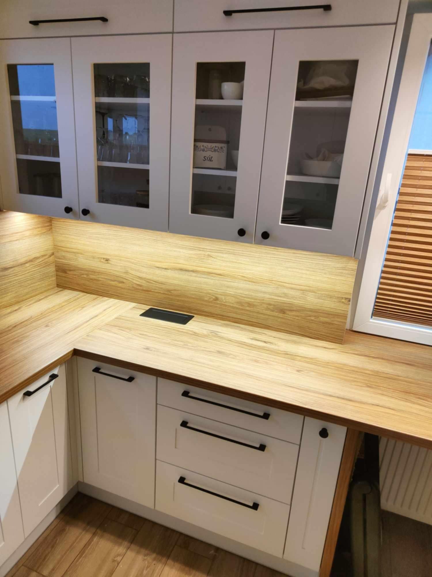 Jasna kuchnia z drewnianym blatem i szafkami z przeszkleniami. Nowoczesne czarne uchwyty. Widoczny fragment okna z drewnianymi żaluzjami.