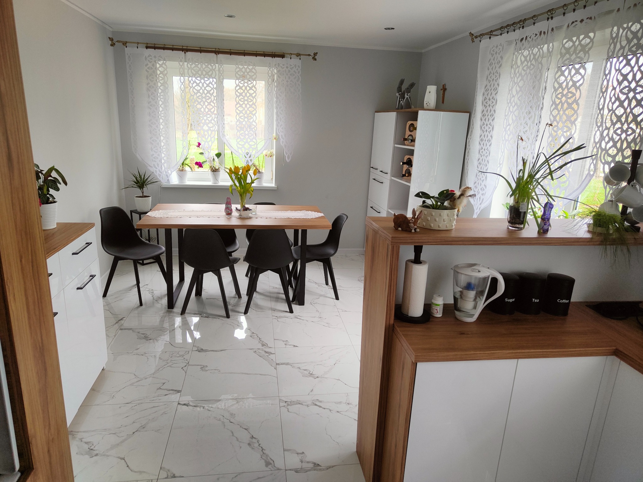 Jasna jadalnia z drewnianym stołem i czarnymi krzesłami na tle marmurowej podłogi. Białe szafki z czarnymi uchwytami, okno z koronkowymi firankami i rośliny doniczkowe.