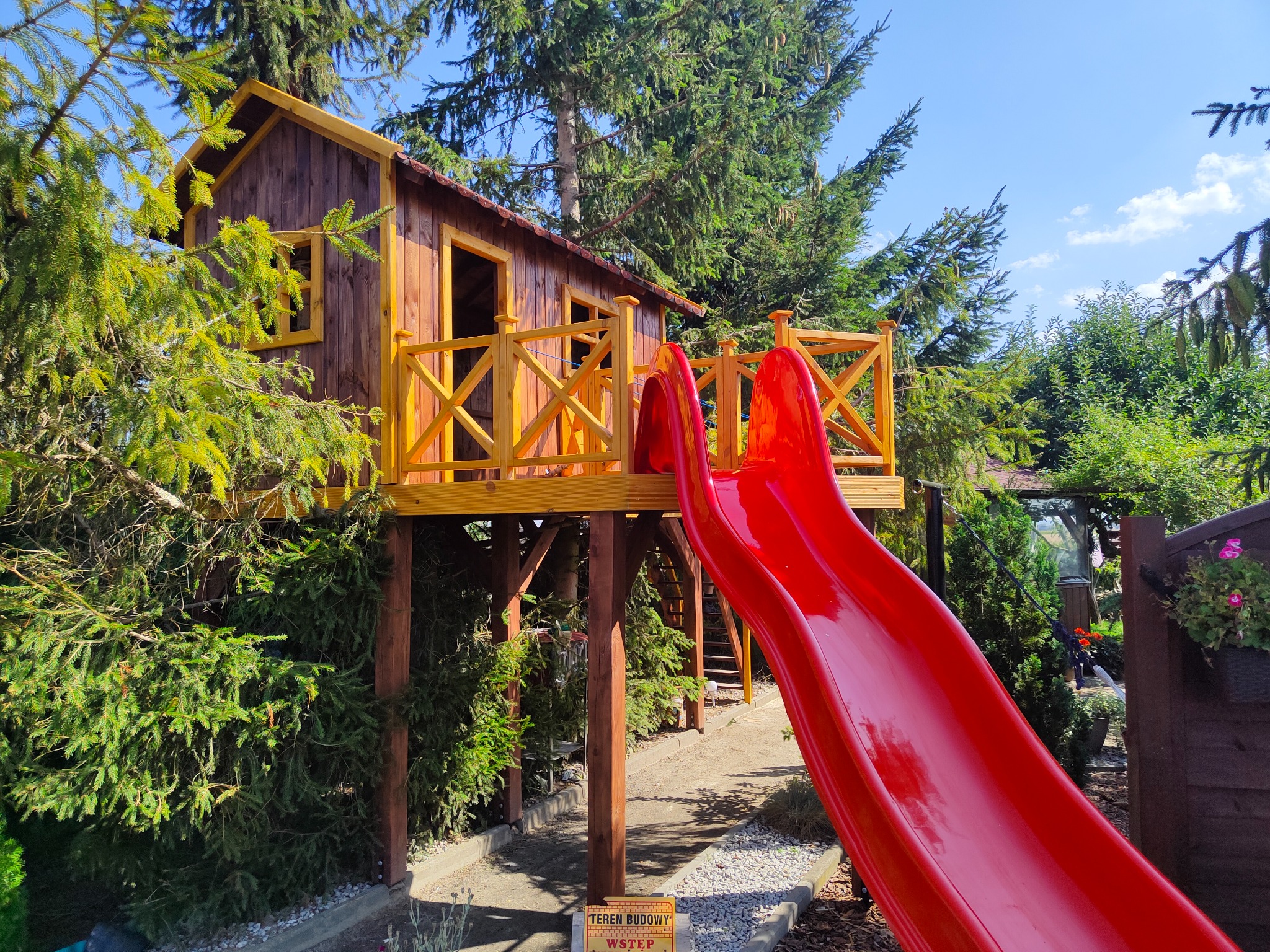 Drewniany domek na drzewie z czerwoną zjeżdżalnią w ogrodzie, otoczony zielenią. Konstrukcja wsparta na palach, z żółtymi balustradami. Tabliczka 'Teren Budowy' w tle.