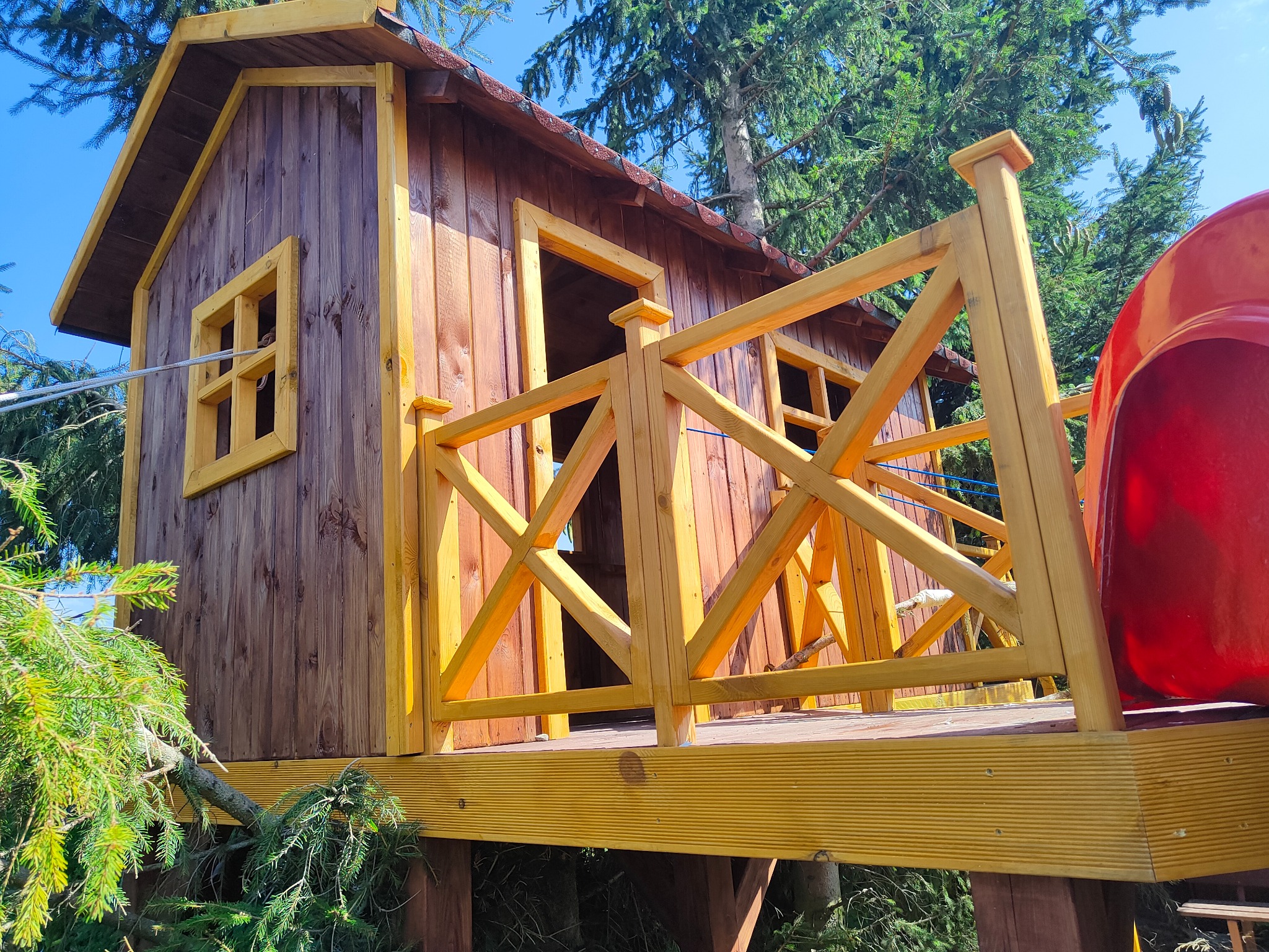 Drewniany domek na drzewie z żółtą balustradą i czerwoną zjeżdżalnią, otoczony zielenią drzew, widziany z dołu pod kątem, w słoneczny dzień.