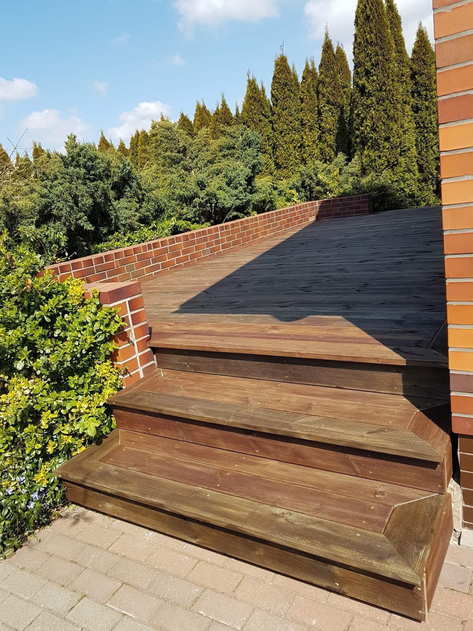 Drewniany taras z trzema schodkami, otoczony zielenią i ceglanym murem, w słoneczny dzień. Widoczne deski tarasowe i elementy konstrukcyjne.