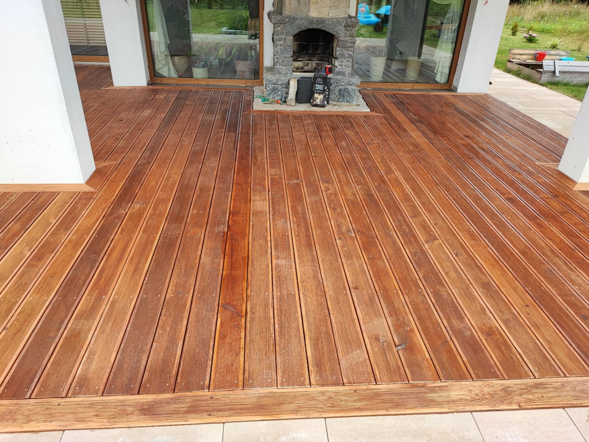 Wykończony taras z desek ryflowanych w odcieniach brązu, z paleniskiem z kamienia. Widok z poziomu podłogi, podkreślający strukturę drewna i solidne wykonanie.