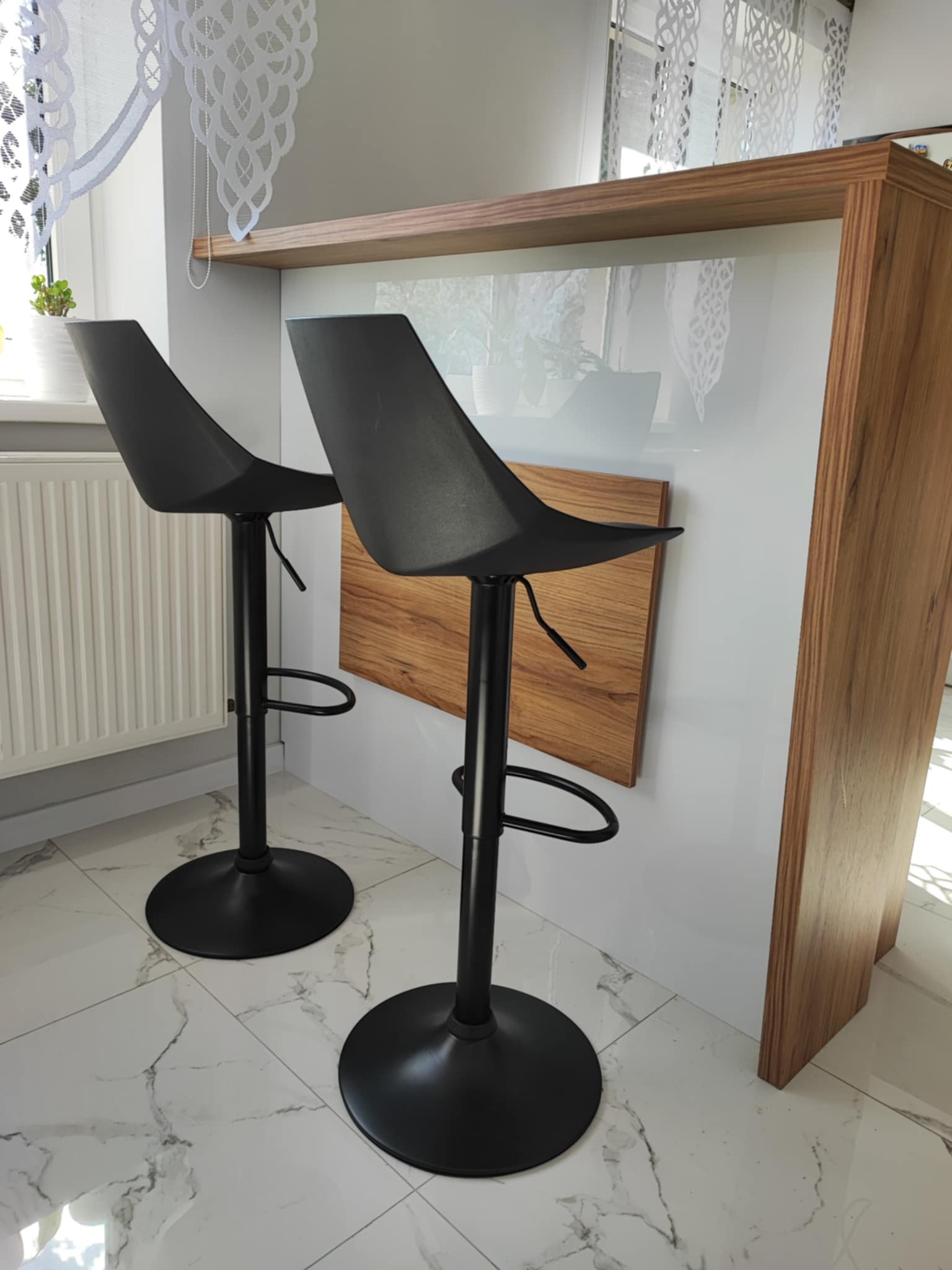 Dwa czarne hokery przy drewnianym blacie z białym frontem. Widoczny fragment okna z firanką i kaloryfer. Marmurowe płytki podłogowe.