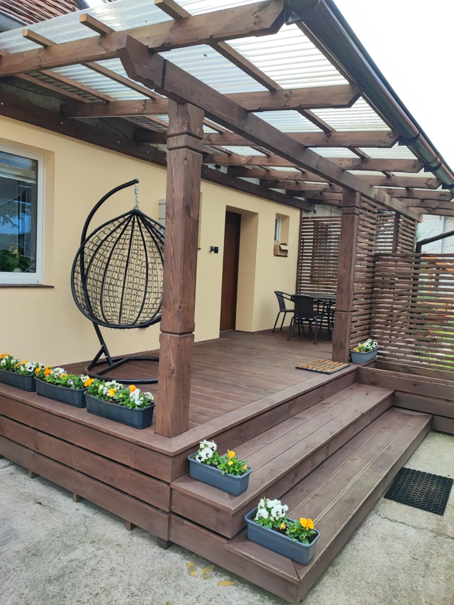 Zadaszony taras z drewna z huśtawką typu kokon i kwiatami w donicach. Widoczny stół z krzesłami i drewniane ażurowe ogrodzenie.
