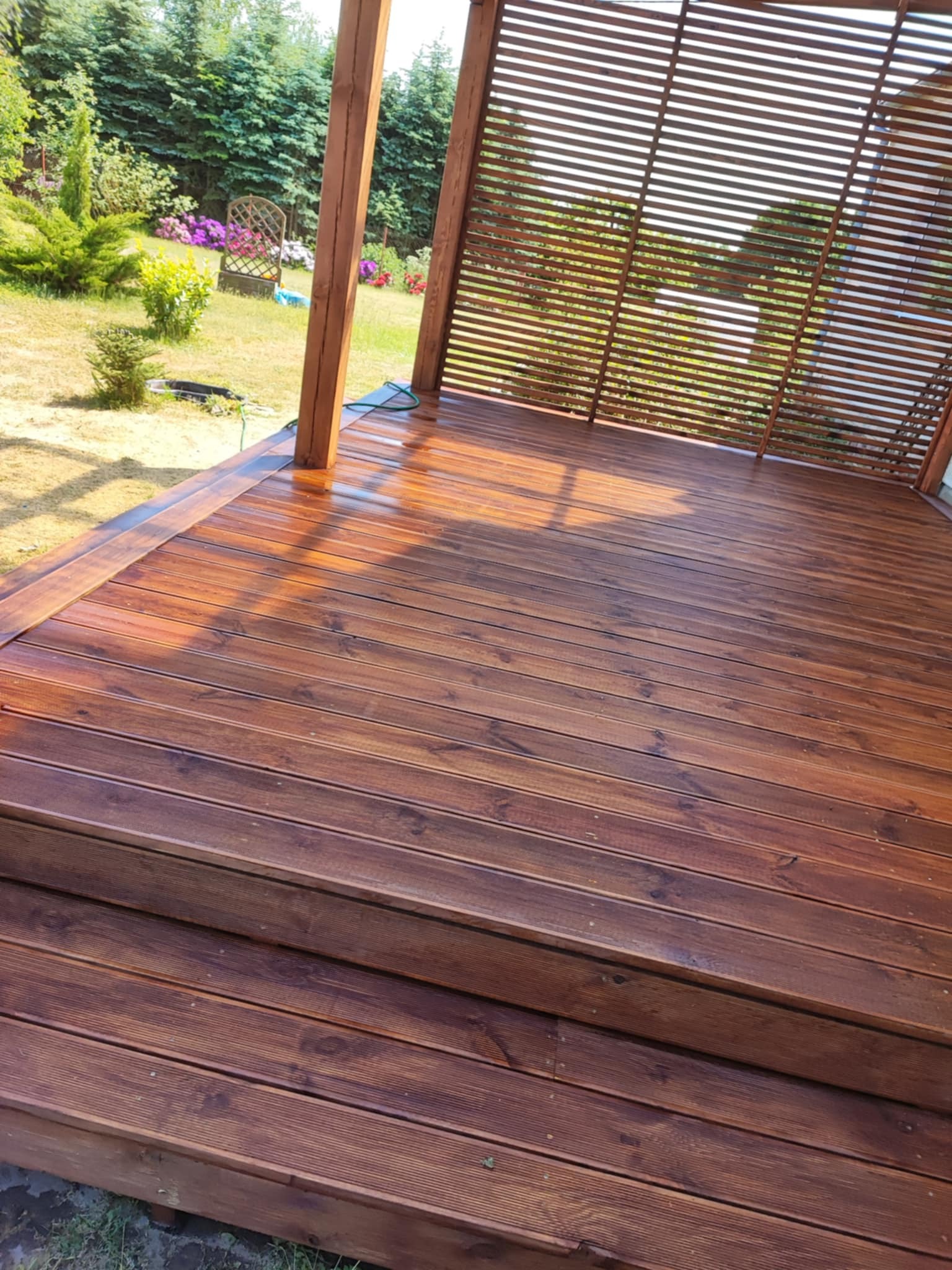 Drewniany taras z zadaszeniem lamelowym w Dąbrówce, wykończony w ciepłym, brązowym odcieniu. Widoczne schody i otaczający ogród.