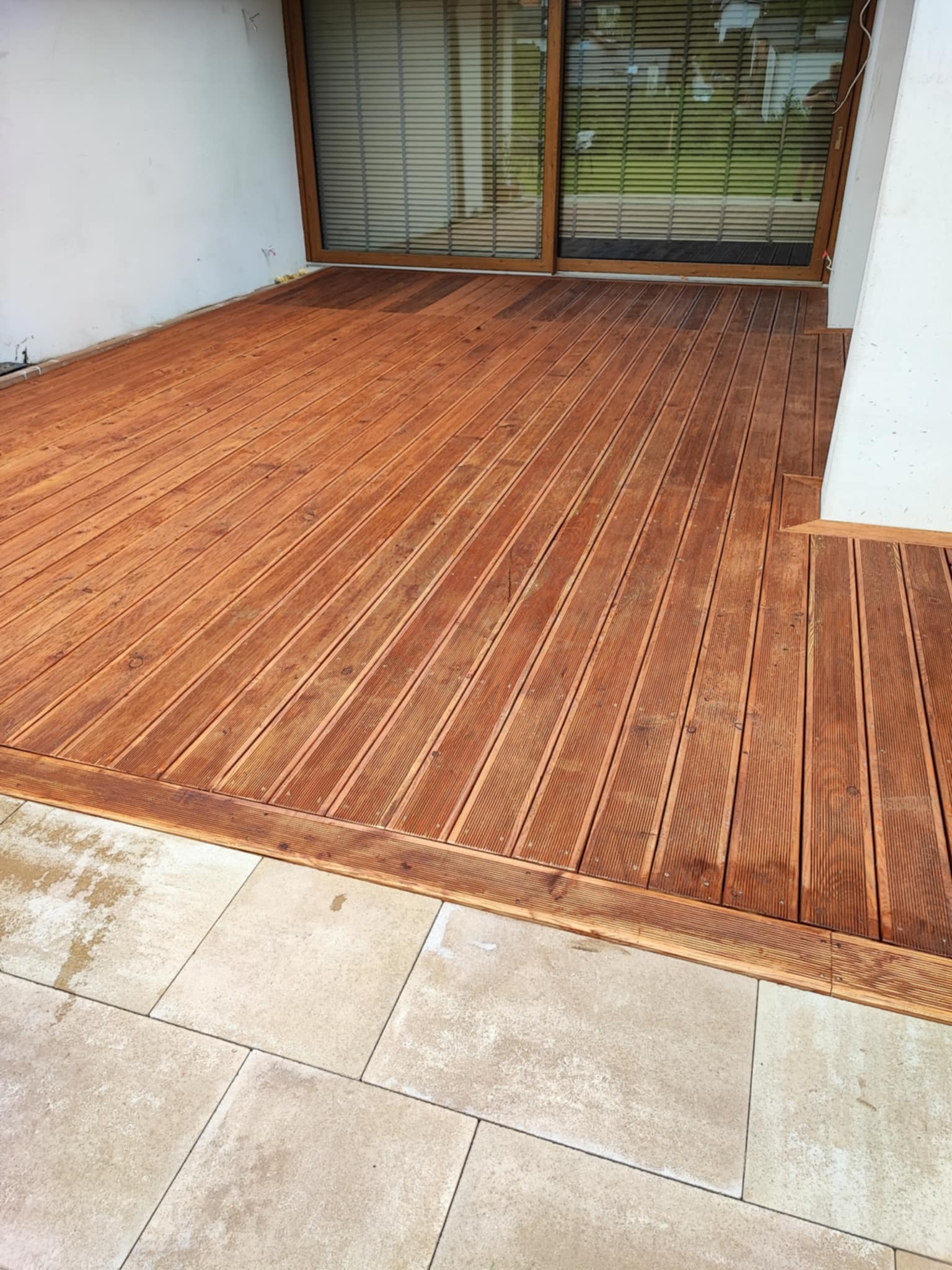 Rustykalny taras drewniany z desek ułożonych pod kątem, z obrzeżem i widoczną kostką brukową oraz oknem tarasowym w tle.