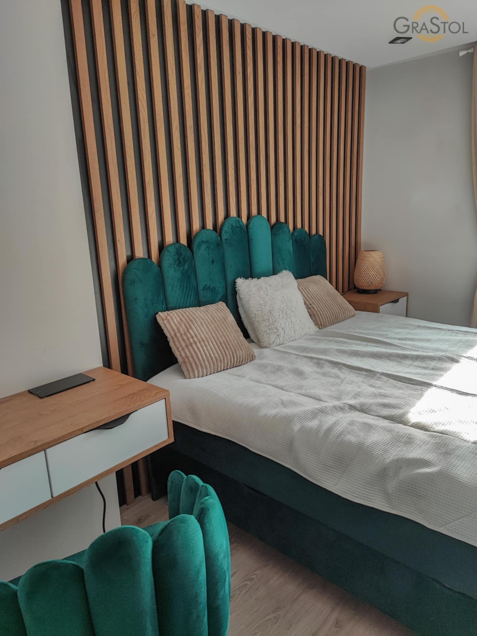 Nowoczesna sypialnia z zielonym, aksamitnym łóżkiem i drewnianą ścianą z lameli. Na szafce nocnej stoi lampka. Widoczny fragment zielonego krzesła.