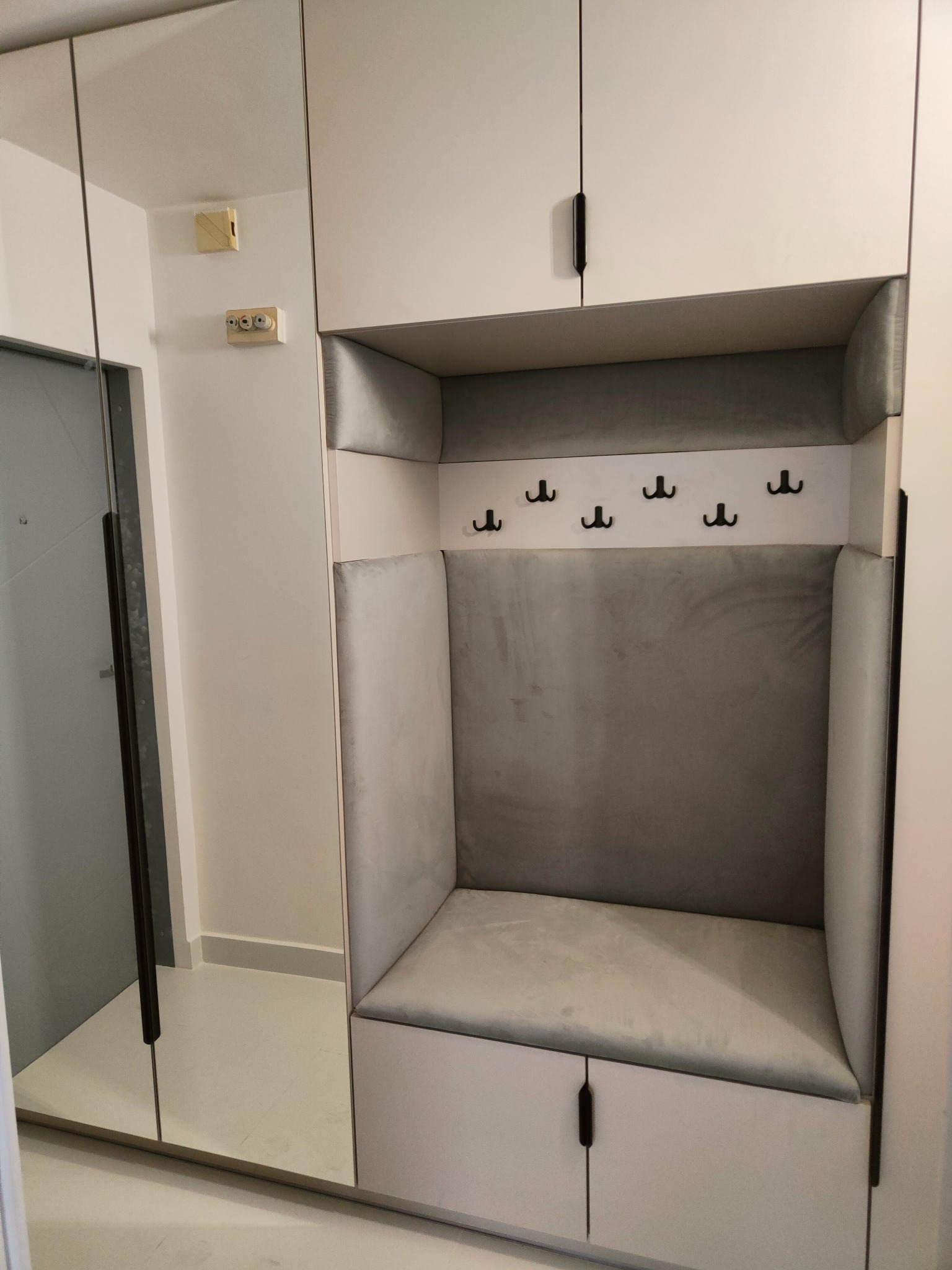 Funkcjonalna zabudowa przedpokoju z lustrem i tapicerowanym siedziskiem z wieszakami. Minimalistyczny design, jasna kolorystyka, czarne uchwyty. Widoczny fragment instalacji elektrycznej.