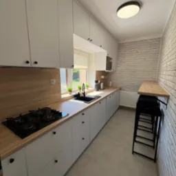 Jasna, nowoczesna kuchnia z białymi szafkami, drewnianym blatem i ceglaną ścianą. Widoczny zlew, płyta gazowa i wysoki stół z krzesłami barowymi.