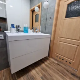 Nowoczesna łazienka z białą szafką pod umywalkę, drewnianą podłogą i drzwiami. Widoczne oświetlenie punktowe i szara ściana z płytkami. Elementy armatury.